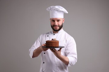 Happy confectioner in uniform holding delicious chocolate cake on light grey background