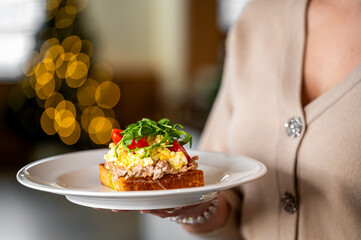 A gourmet open-faced sandwich with scrambled eggs, tuna, cherry tomatoes, and arugula on a white plate. The festive bokeh lights in the background suggest a holiday or celebratory atmosphere