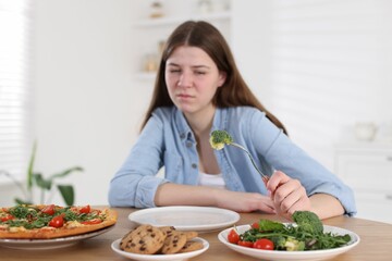 Sad teen girl holding fork with broccoli at home, selective focus. Eating disorder