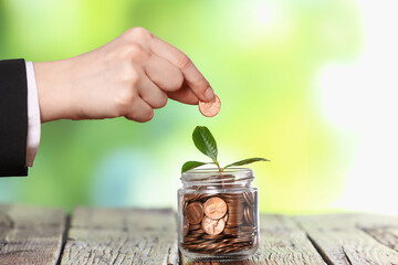 Investment concept. Woman putting coin into glass jar at wooden table against blurred green...