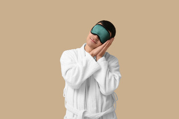 Portrait of sleepy young man in bathrobe and sleeping mask on beige background