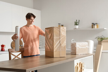 Young man wrapping cardboard box with stretch film on table in kitchen