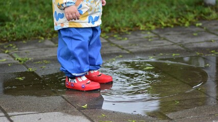 Toddler in colorful rain gear playing in a puddle on a wet sidewalk. Child enjoying rainy day...