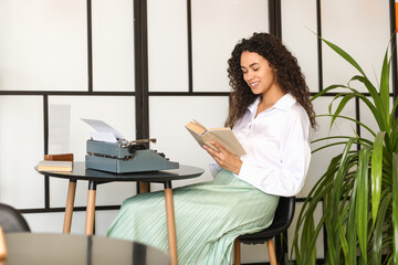 Young African-American author with typewriter reading book at table in cafe