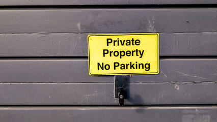 Bright yellow "Private Property No Parking" sign on a dark, weathered garage door, clearly indicating restricted access and parking regulations