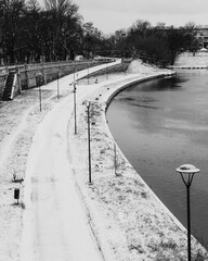 Snow covered promenade lining the river in winter, featuring street lamps, benches, and trash cans,...