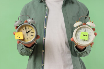 Man holding alarm clocks with word TIME and paper smile on green background, closeup