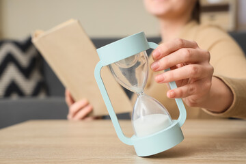 Woman with hourglass reading book at home, closeup