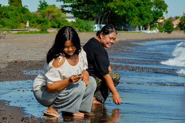 A teenage girl squats to pick up a small stone on the shore, focused on the ground with gentle...