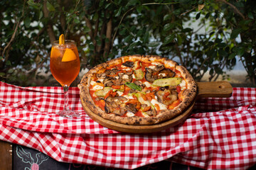 Wood-fired vegetable pizza and orange cocktail on checkered picnic cloth.
