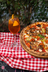 Wood-fired vegetable pizza and orange cocktail on checkered picnic cloth.
