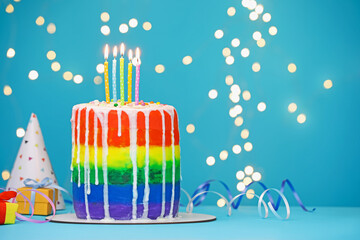 Delicious rainbow birthday cake with burning candles, streamers, gift boxes and party hat on light blue table against blurred lights, closeup. Space for text