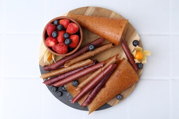 Delicious fruit leather rolls and berries on white tiled table, top view