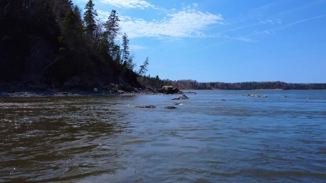 A low-level flight across the mouth of a fast-flowing river to its bank, which is made up of rocky slumps surrounded by forest, under a blue sky. Mitis River, Quebec, Canada, 2025.
