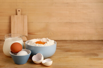 Bowl of flour with fresh chicken eggs and jug of milk on wooden table