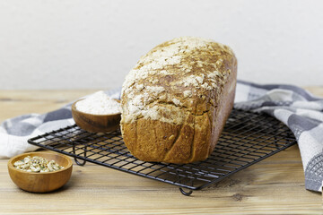 A loaf of bread rests on a cooling rack on a wooden table. The bread is made with whole-grain rye flour and has a crispy crust. Next to the bread is a bowl of flour and seeds.