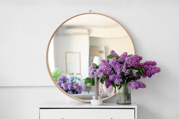 Vase with beautiful lilac flowers and candle on chest of drawers in room