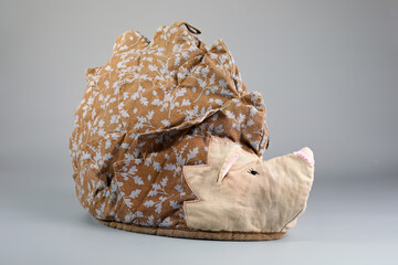 A whimsical hedgehog-shaped tea cosy, crafted from patterned fabric. The brown body features a white leaf print, complemented by a light-coloured, quilted face. Decorative and functional.
