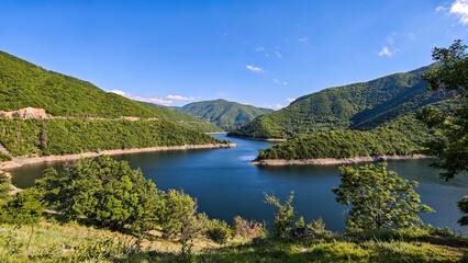 Panoramic view of Vacha Reservoir in Bulgaria, surrounded by lush green mountains and calm blue water under a bright sky.