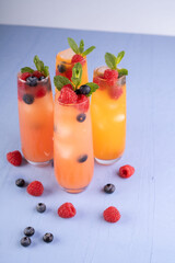 vibrant orange and red berry infused summer cocktails with mint garnish in tall glasses on light blue textured surface complemented by scattered fruits, trendy, fashionable, tempting, delightful
