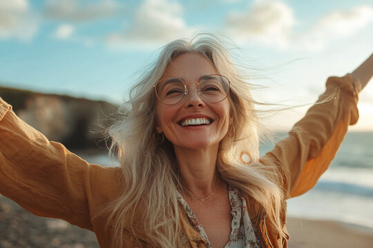 Joyful mature woman in a serene beach setting at sunset, embracing life's beauty with open arms and a beaming smile, reflecting happiness, peace, and fulfillment.