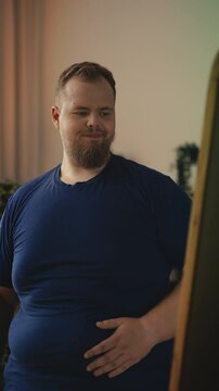 Overweight man looking at his reflection, holding belly, unhappy with body image