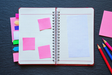 Open spiral bound notebook with blank pages, notes and colorful bookmarks. Colored pencils on black background. Copy space