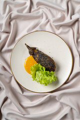 A beautifully arranged plate showcases a perfectly grilled eggplant resting beside a bright lemon sauce and fresh lettuce. The soft pink fabric creates a delicate backdrop for this culinary delight