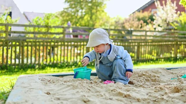 Two-year-old boy in a Panama hat on his head plays in a sandbox with sand on a playground. Toddler in stylish casual clothes digs sand while sitting in a sandbox on the street. Child development.