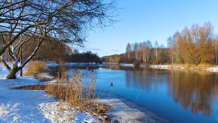 The river flows through a mixed forest. There are ripples on the water and reeds near the shore. In winter there is snow on the ground and tree branches. Sunny and frosty weather and a blue sky