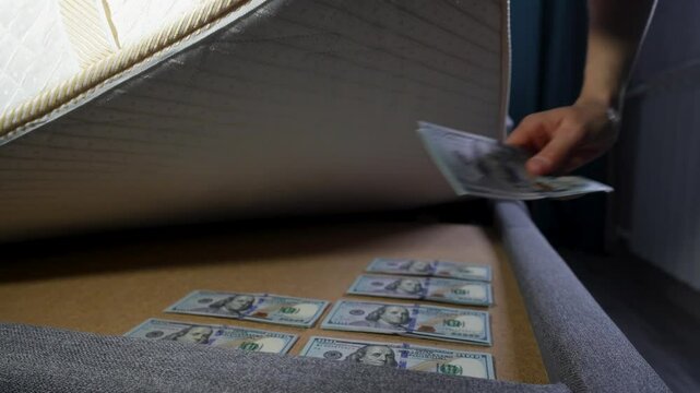Close-up of a person taking a stack of US hundred-dollar bills under a mattress