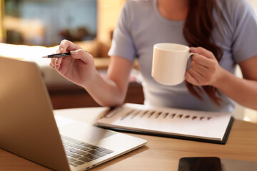 Asian women hand holding pen and coffee cup working with laptop and graph for work from home...
