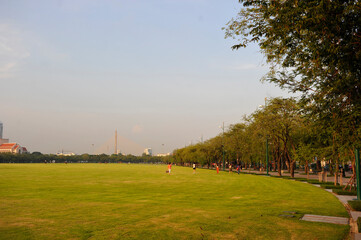 A large open green park with a few people walking and trees lining the sides under a clear sky. Bangkok Thailand.