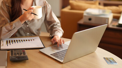 Asian woman analyzes charts on laptop with coffee cup in her hand Financial Planning and Investment...