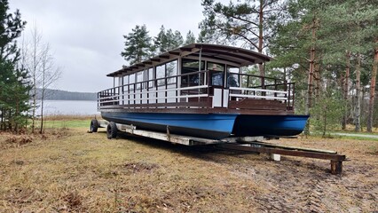 On the grassy shore of the lake among pine trees stands a carriage, and on it is placed a catamaran with a passenger cabin for tourist water trips. A mixed forest grows around the lake. Overcast