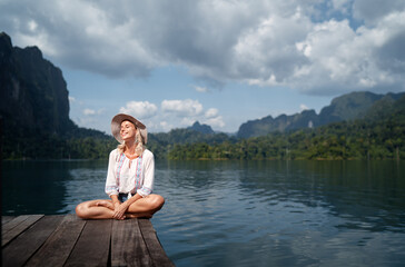 Fototapeta premium A serene moment as a woman enjoys the tranquil lake surroundings amidst beautiful mountain scenery. She sits relaxed on the wooden dock, basking under the sun with a cheerful expression.
