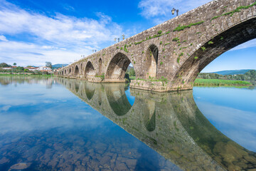 Fototapeta premium The historic Ponte de Lima bridge over perfectly still water in Portugal, with the San Antonio Church reflected beautifully under a clear blue sky.