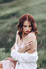 A beautiful red-haired young woman in a pink dress is sitting in nature among flowers at sunset. In the background, there are hills and ravines. Close-up portrait