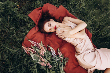 A beautiful red-haired young woman in a pink dress is lying on a red blanket in nature among flowers at sunset. In the background, there are hills and ravines.