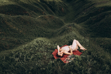A beautiful red-haired young woman in a pink dress is lying on a red blanket in nature among flowers at sunset. In the background, there are hills and ravines.