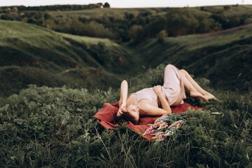 A beautiful red-haired young woman in a pink dress is lying on a red blanket in nature among flowers. In the background, there are hills and ravines. Focus on the reed. The girl is out of focus