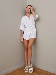 Fotobehang A woman stands against a corner wall with a confident pose, wearing a tied white shirt and shorts set. She has her hand touching her head, sandals on, wooden floor underfoot © Dmitry Tsvetkov