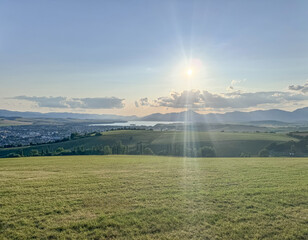This stunning landscape features rolling hills, a serene lake, and majestic mountains, illuminated by the warm glow of the setting sun and soft rays of light breaking through clouds