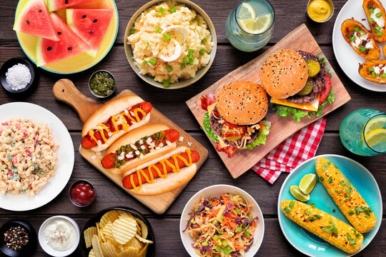 Summer food table scene with hot dogs, hamburger and sides. Overhead view over a dark wood background.