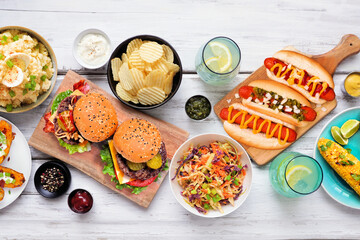 Summer food table scene with hot dogs, hamburger and sides. Above view over a white wood background.