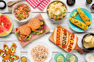 Summer food table scene with hot dogs, hamburger and sides. Top down view over a white wood background.