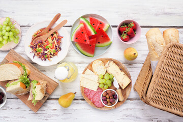 Summer picnic food table scene. Top view over a white wood background. Picnic basket, fruit, salad, sandwiches and snacks.