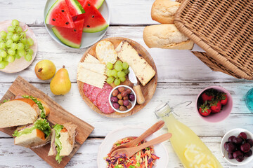 Summer picnic food table scene. Top view over a white wood background. Picnic basket, fruit, salad, sandwiches and snacks.