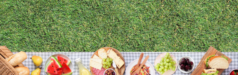 Summer picnic food bottom border. Top view with blanket over a green lawn banner background. Picnic basket, fruit, salad, sandwiches and snacks. Copy space.