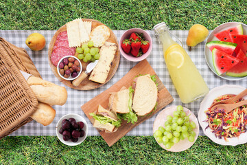 Summer picnic food scene. Top view with blanket over a green lawn background. Picnic basket, fruit, salad, sandwiches and snacks.